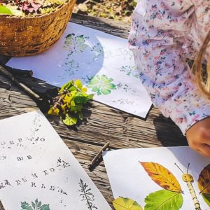 Un cuaderno de naturaleza abierto sobre una mesa de madera, con hojas y flores estampadas en sus páginas, junto a una cesta de hojas y unas manos infantiles colocando una margarita amarilla